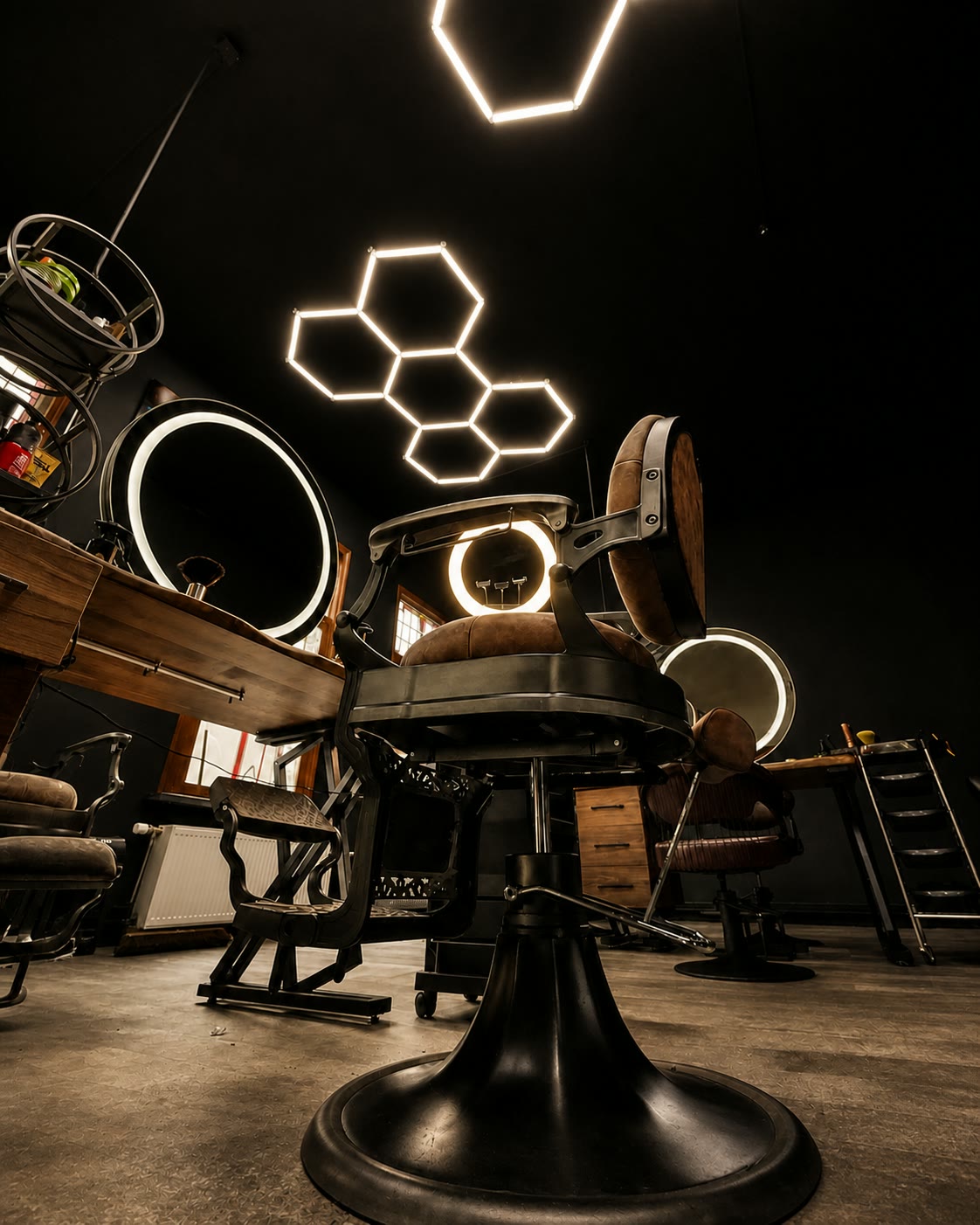 The empty shop at rest: cast-iron barber chairs, a round ring-light mirror and hexagon neon panels across the black ceiling.