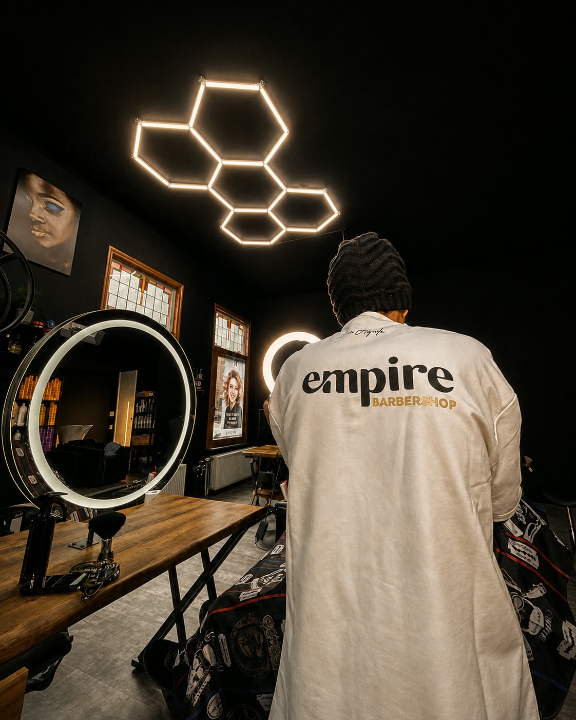 A barber in an empire BARBERSHOP tee stands with his back to camera in the long mirrored room.