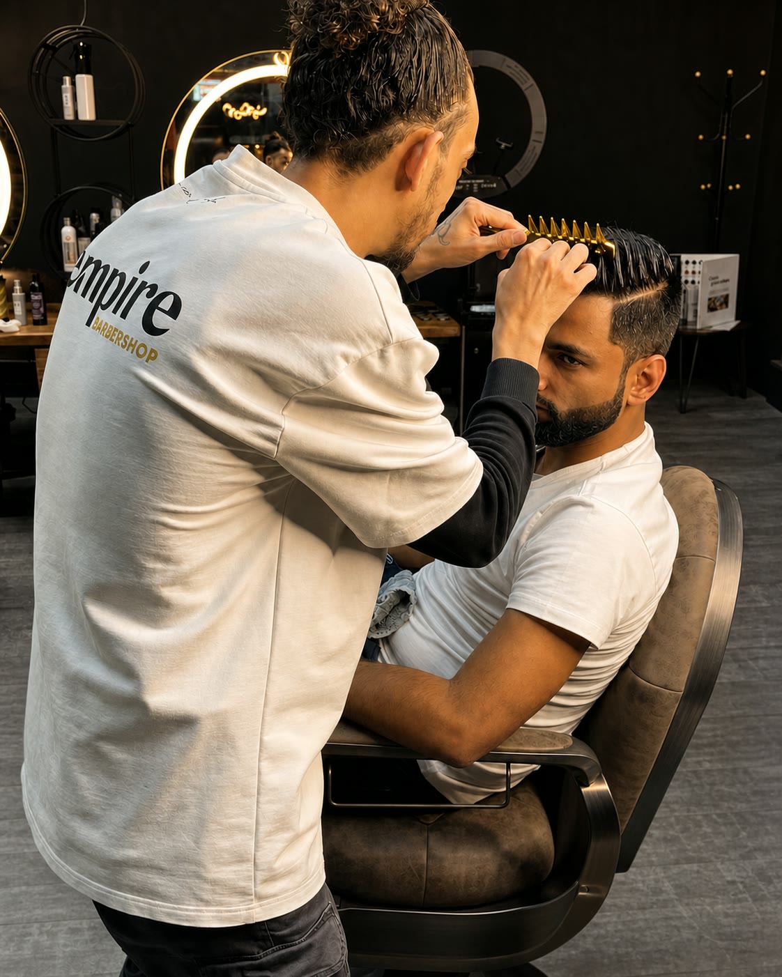 A barber leans in with clippers to work the hairline of a seated client, both of them in white tees.
