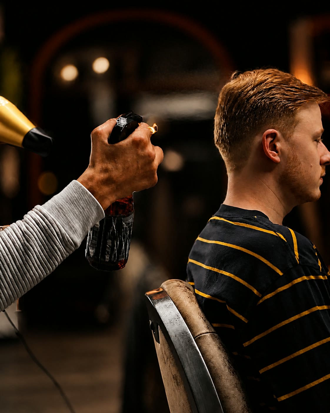 A barber finishes the same red-haired client with a dryer and a brush, the client seen in profile.