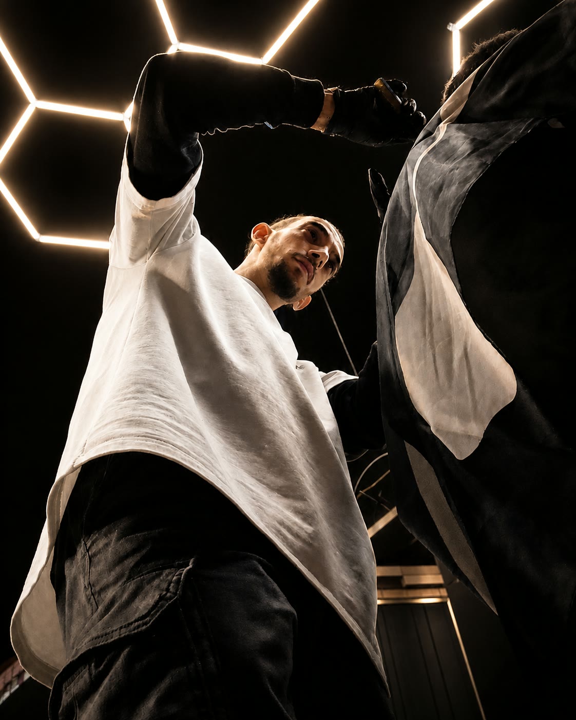 A barber sweeps the cape off a client in one movement, lit from below against the black wall and the hexagon neon.
