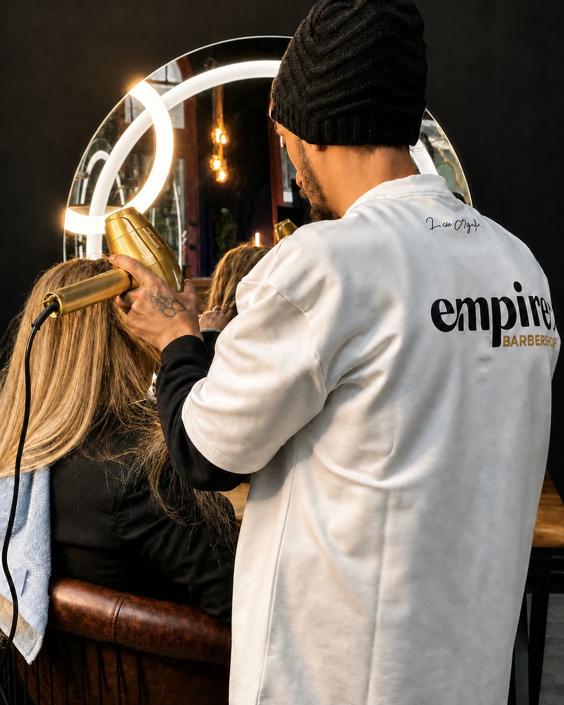 A barber in a black beanie blow-dries long blonde hair, seen from behind under the ring light.